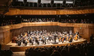 Orkiestra symfoniczna grająca w nowoczesnej sali koncertowej, z widownią na pierwszym planie.